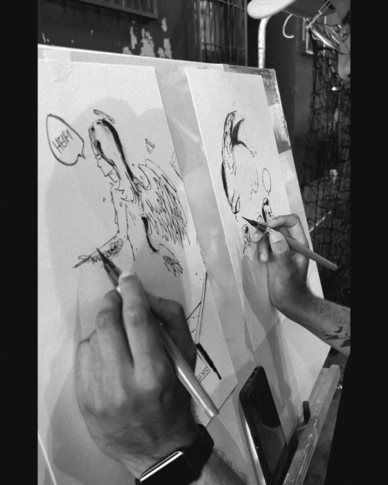 Gros plan en noir et blanc des deux mains d’Isia dessinant simultanément sur deux feuilles.